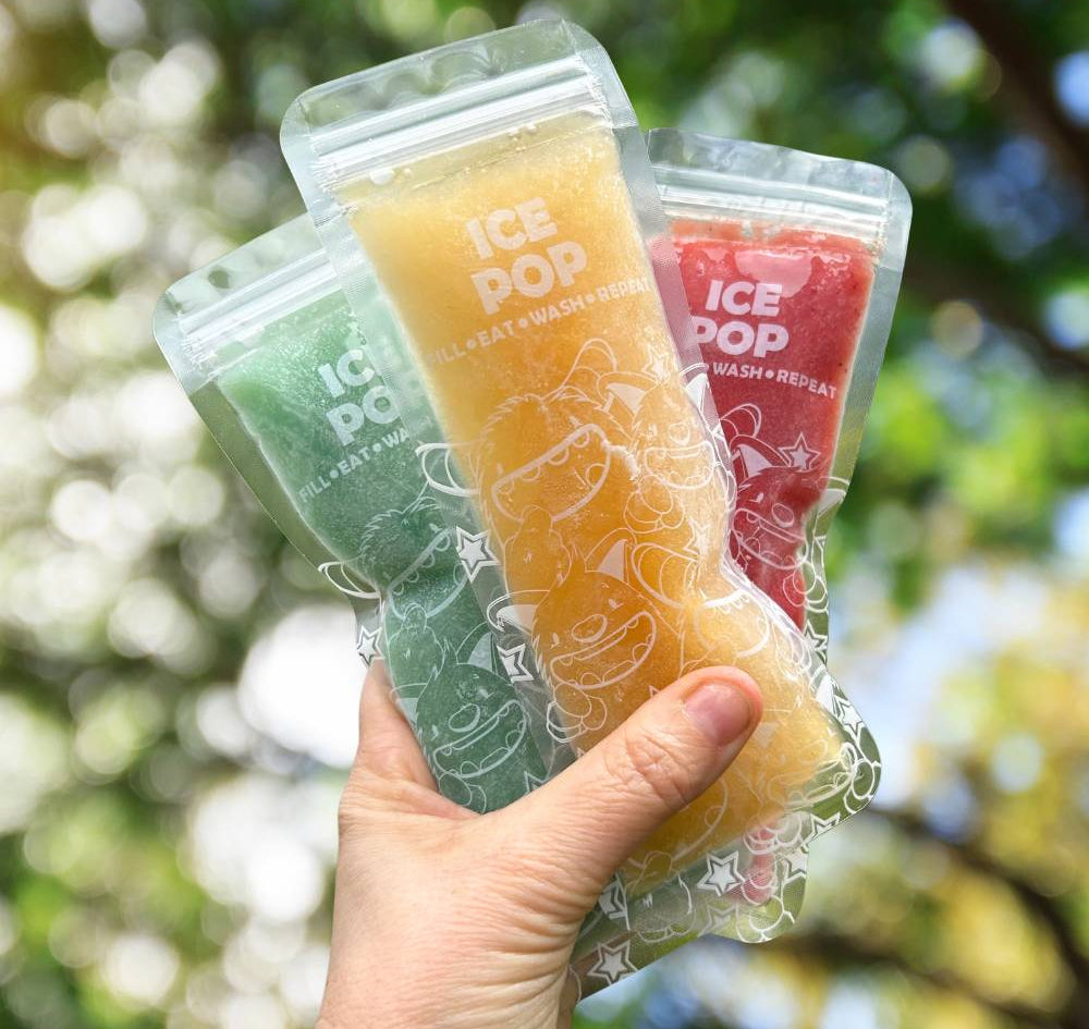 Hand holding three ice pop packages with colorful popsicles against a blurred green outdoor background