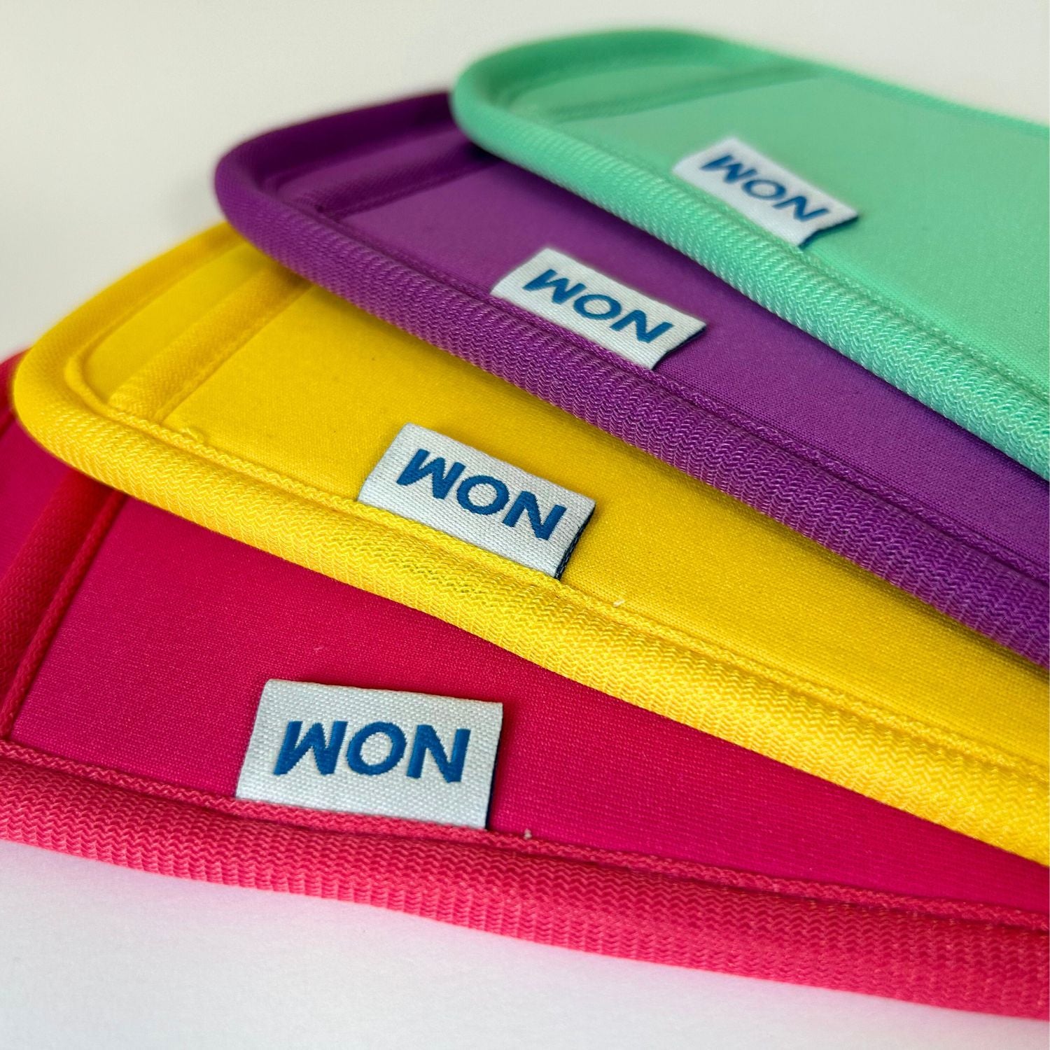 A set of four neoprene ice pop holders in pink, purple, yellow, and teal, each with a label showing the brand name 'Nom Nom'.