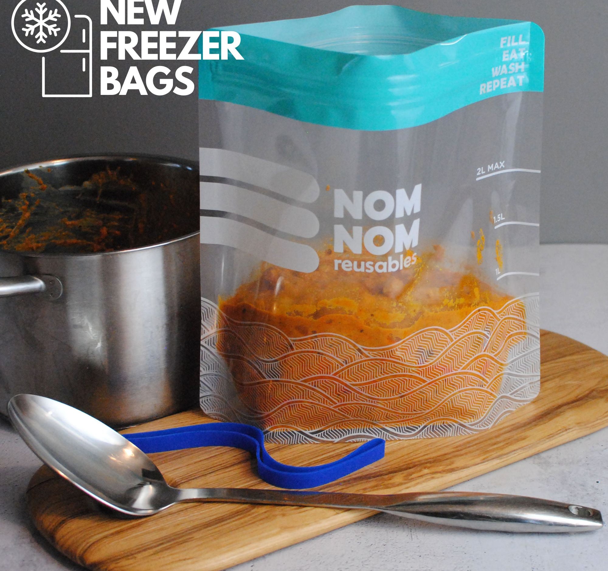 A Nom Nom clear reusable freezer bag filled with food, alongside a silver spoon on a wooden board.