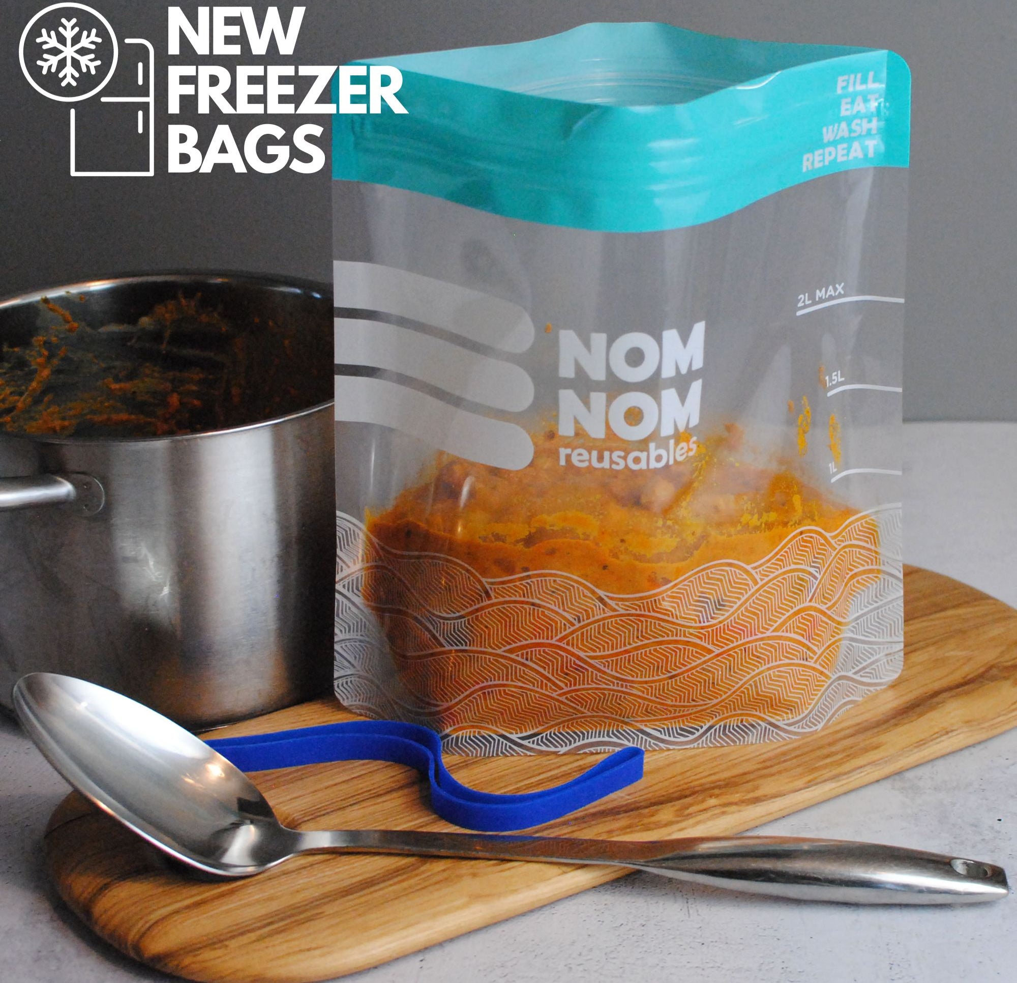 A Nom Nom clear reusable freezer bag filled with food, alongside a silver spoon on a wooden board.