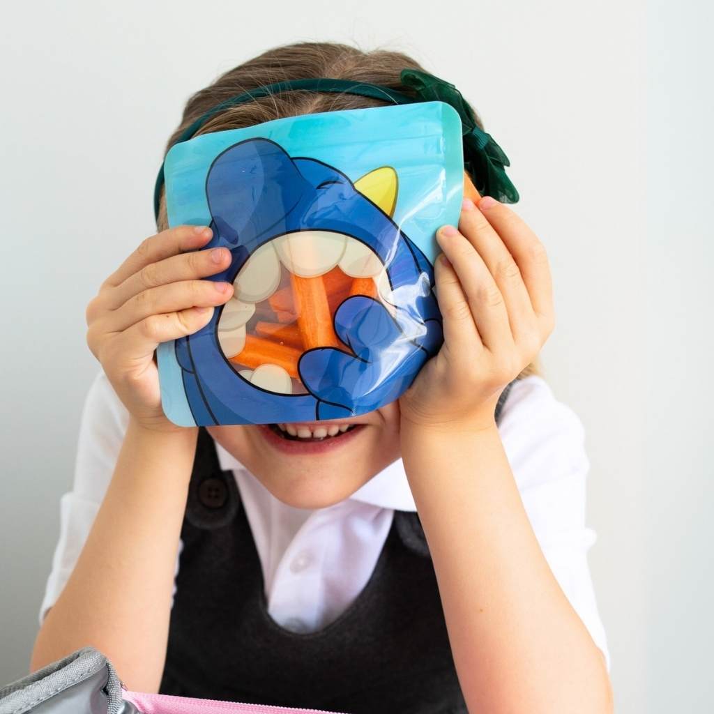 child holding a blue monster reusable snack bag against her face, The bag is filled with chopped carrots for her snack
