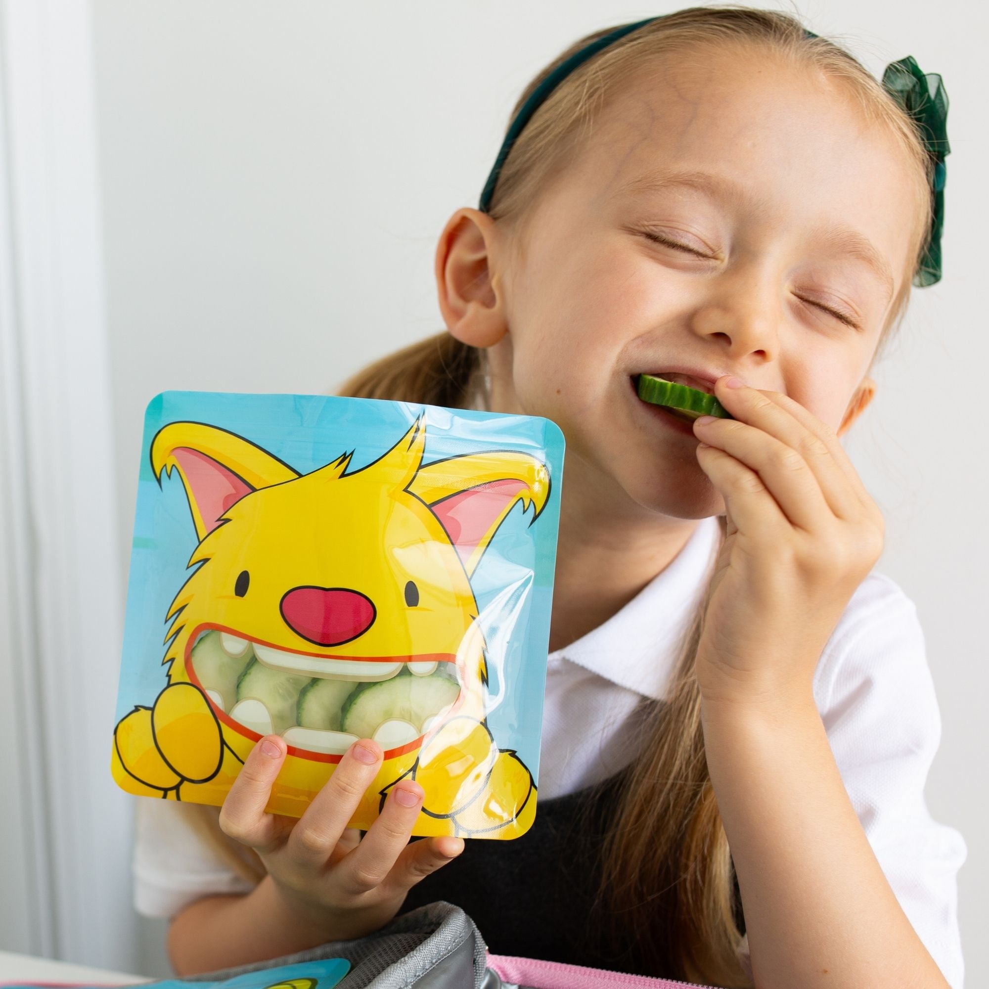 reception age school girl sat at lunch time eating cucumber from a yellow monster snack bag