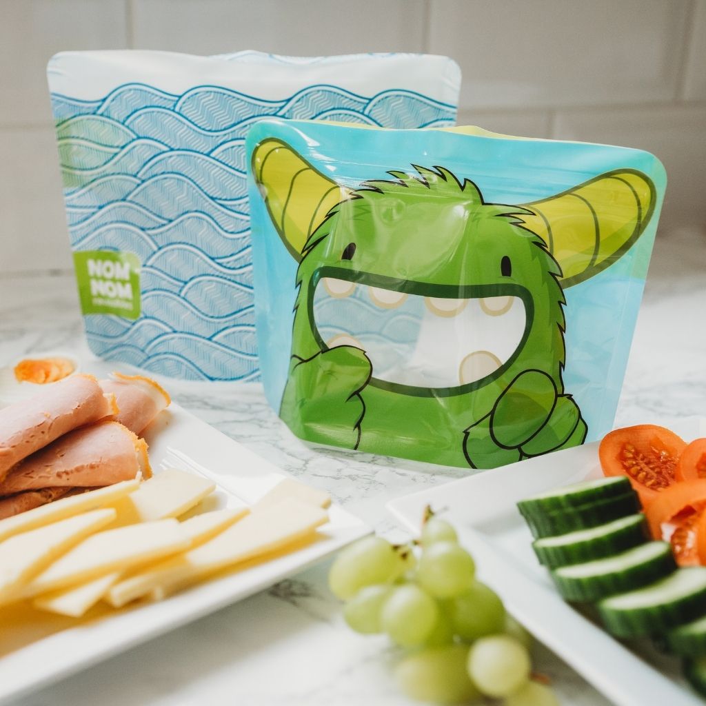 Green monster reusable sandwich bag on a table with an adults wave design sandwich bag ready to be filled for lunch with a selection of bread, cheese, ham, grapes, and tomatoes