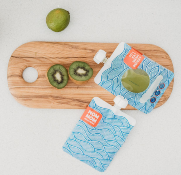 Green smoothie in a large capacity reusable pouch from the Nom Nom Kids wave range for older children, teens and adults. The pouches are on a chopping board with a kiwi and lime.