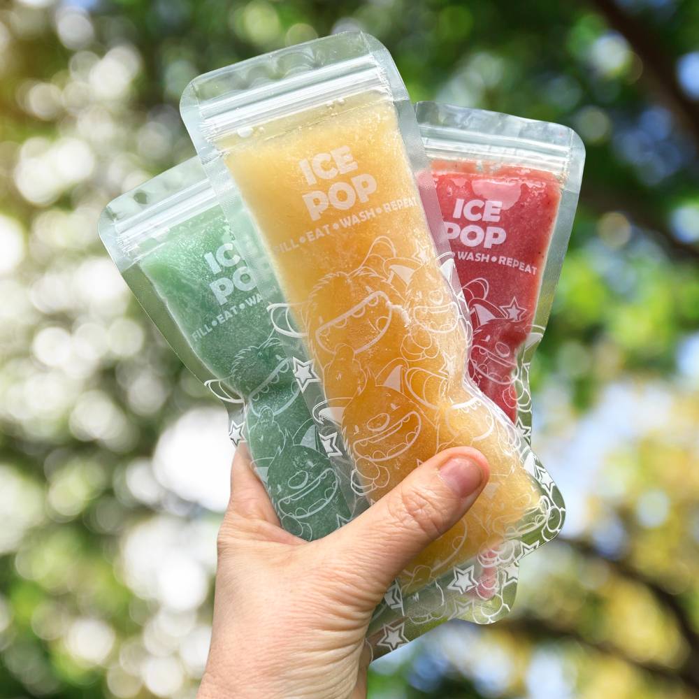 Hand holding three ice pop packages with colorful popsicles against a blurred green outdoor background