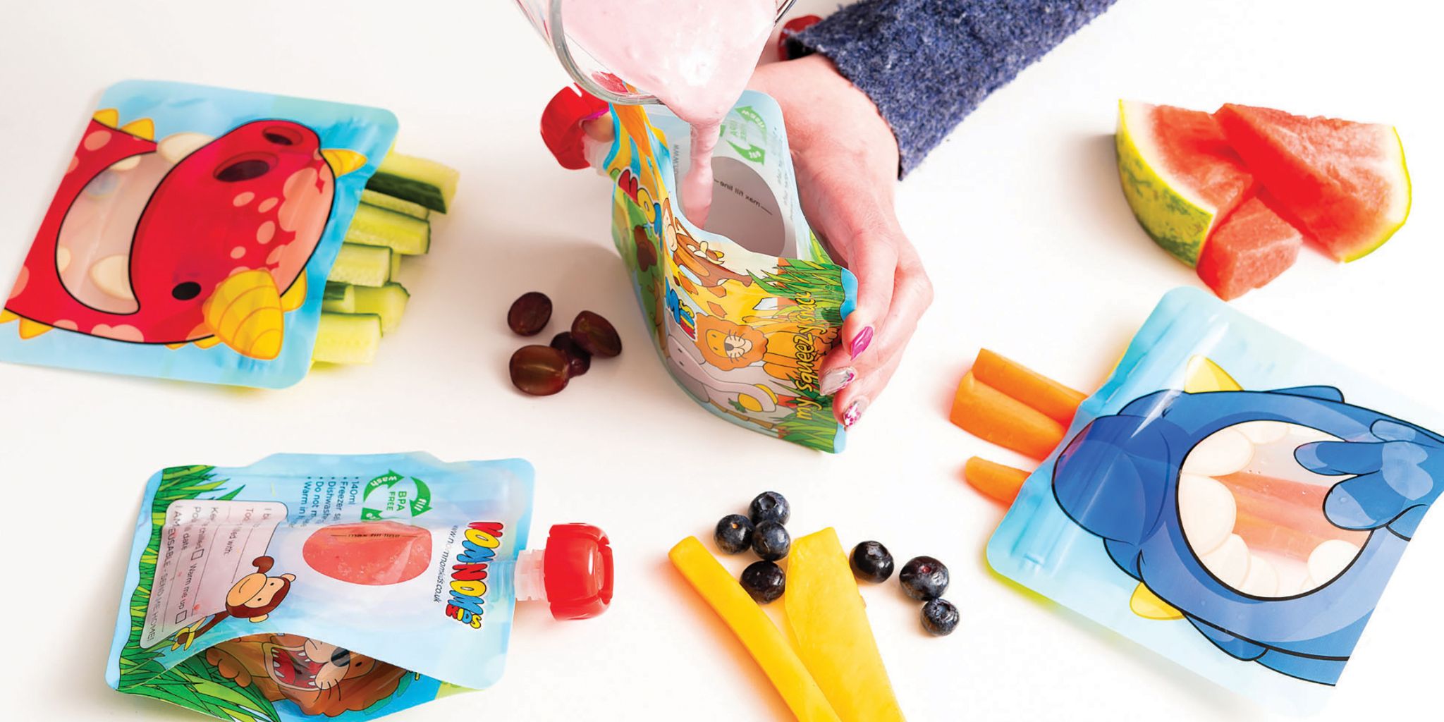 A nom nom kids reusable pouch being held open in one hand and a jug of yogurt smoothie being poured in with the other. Surrounded by snack bags and a filled pouch.