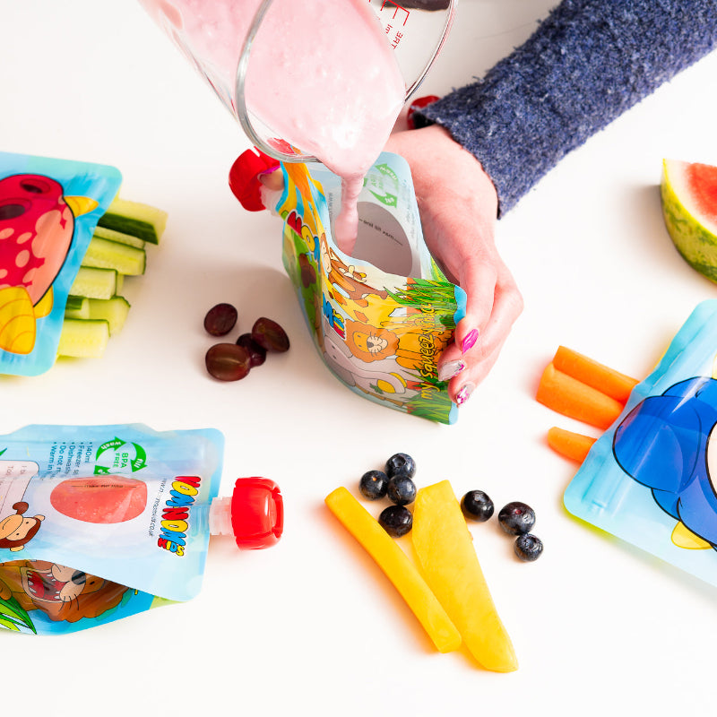 A nom nom kids reusable pouch being held open in one hand and a jug of yogurt smoothie being poured in with the other. Surrounded by snack bags and a filled pouch.