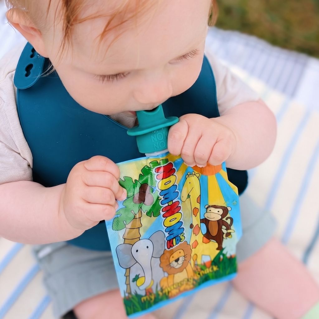 Weaning baby holding a Nom Nom Kids pouch with animal illustrations and topped with a silicone pouch spoon on blue